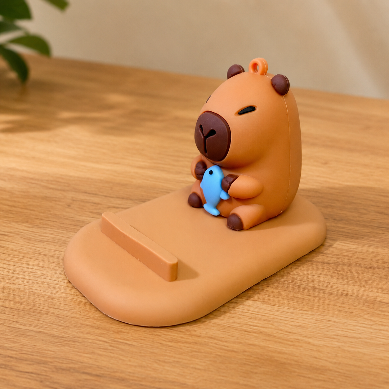 capybara phone stand holding a blue fish on a wooden stand with a plant in the background
