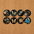 Set of eight decorative coasters with floral and butterfly designs on a wooden surface.