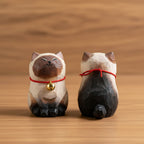 Two wooden cat figurines with red necklaces and a gold bell on a stone ledge with green foliage in the background.