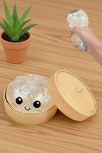 Small round squishy toy with a face on a wooden surface, next to an open beige container.