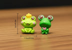 Two small green frog figurines with measurements on a gray surface.