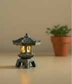 Decorative japanese style stone lantern with glowing interior on a wooden surface, potted plant in the background.