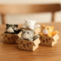 4 Cute Cat figures of various colours in decorative cardboard boxes on a wooden surface.